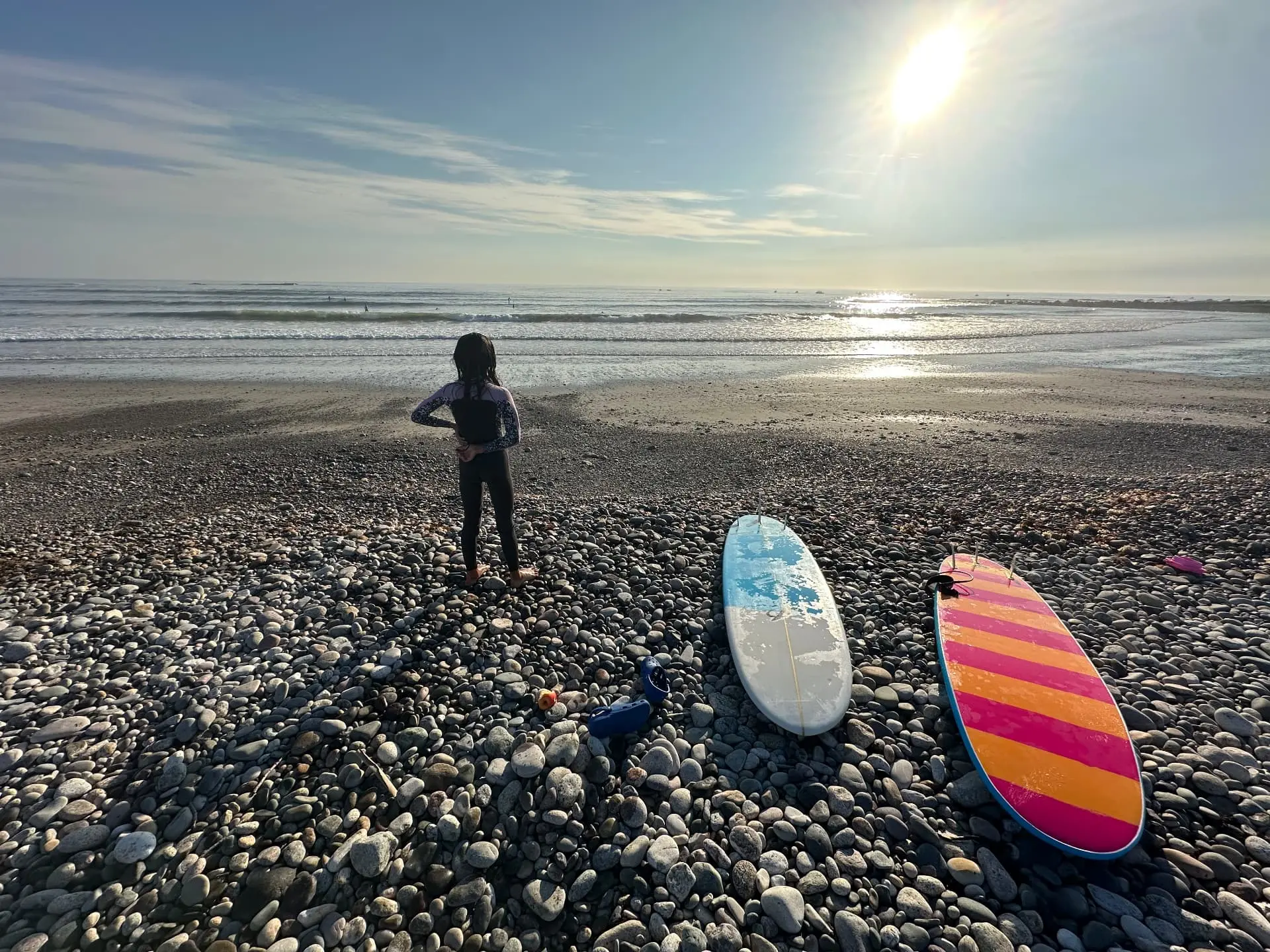 Girl in a wetsuit on a pebble beach at sunrise beside two surfboards