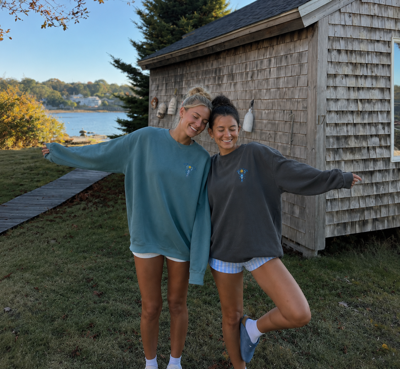 Two women in Checkered Lobster crewnecks outside a shingled boathouse on the harbor